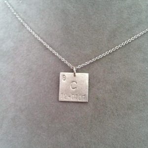 Könnte beinhalten: Eine silberne Halskette mit einem quadratischen Anhänger, der das chemische Symbol für Kohlenstoff, 'C', mit der Ordnungszahl '6' und dem Atomgewicht '12,0107' zeigt.