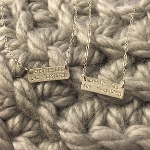 May include: Two silver necklaces with the phrase "Nevertheless, she persisted" engraved on rectangular tags.