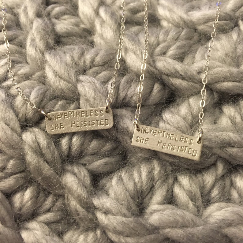 May include: Two silver necklaces with the phrase "Nevertheless, she persisted" engraved on rectangular tags.