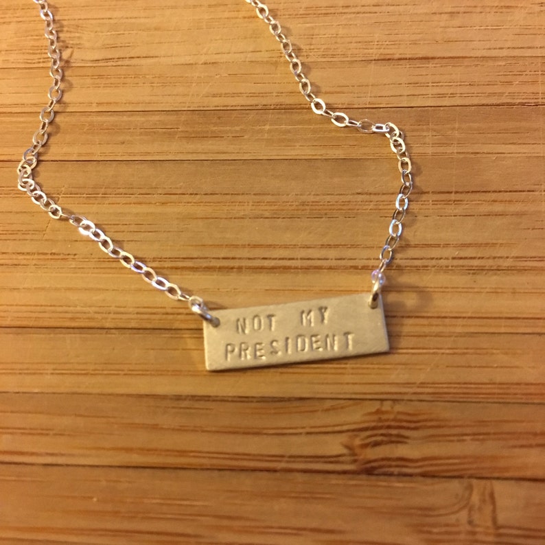 May include: A silver chain necklace with a silver rectangular pendant that says "NOT MY PRESIDENT".