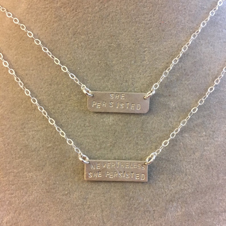 May include: Two silver necklaces with rectangular pendants. The top pendant reads "SHE PERSISTED". The bottom pendant reads "NEVERTHELESS SHE PERSISTED".