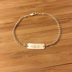 May include: A silver chain bracelet with a rectangular tag that reads "RESIST".