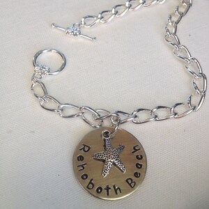 May include: Silver-toned chain bracelet with a toggle clasp. A round, gold-colored charm is engraved with "Rehoboth Beach" and features a starfish. The bracelet is designed for everyday wear and is a beach-themed accessory.