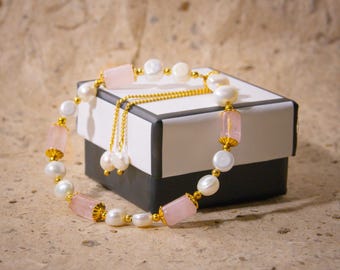 May include: A bracelet featuring white pearls and pink rectangular gemstones, interspersed with gold-colored accents. The bracelet is displayed on a black and white box, with a gold chain and pearl pendant. The jewelry is set against a textured, neutral background.