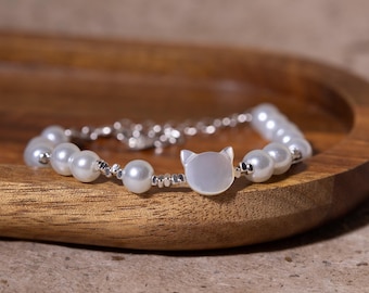 May include: A delicate bracelet featuring faux pearls and a silver-toned chain. The focal point is a cat-shaped charm, adding a whimsical touch. The bracelet is displayed on a wooden tray, showcasing its elegant design.