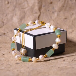 May include: A pearl and jade bracelet with gold detailing sits on a black and white box. The bracelet features white pearls, light green jade stones, and small gold beads. A gold chain with a pearl pendant hangs from the bracelet.