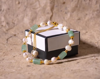 May include: A pearl and jade bracelet with gold accents rests on a black and white box. The bracelet features white pearls, light green jade stones, and small gold beads. A gold chain with a pearl pendant hangs from the bracelet.