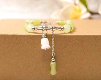 May include: A light green beaded bracelet with a silver-tone bamboo-style center. A white flower charm and a light green teardrop charm dangle from the bracelet. The bracelet is displayed on a brown surface.