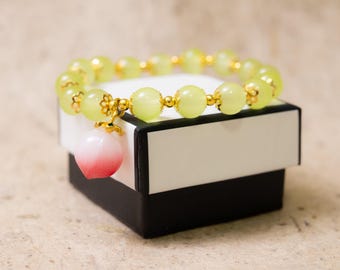 May include: A beaded bracelet with pale green beads and gold accents rests on a white and black box. A peach-shaped charm with pink and red gradients hangs from the bracelet, adding a touch of color and whimsy to the jewelry.