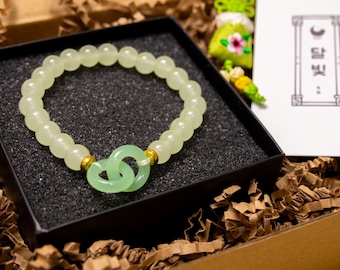 May include: A light green beaded bracelet with a double-ring jade charm, presented in a black box with a textured surface. The bracelet is accented with gold-colored beads. The box sits in a brown cardboard container with shredded paper.