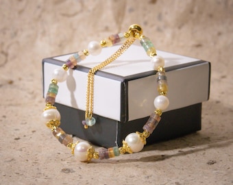 May include: A delicate bracelet featuring a mix of white pearls and colorful, rectangular beads. The bracelet is accented with gold-colored metal and a sliding clasp. The design is set against a neutral background, highlighting the jewelry's details.
