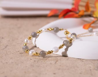 May include: A beaded bracelet featuring gray, white, and gold accents. The bracelet is composed of gray and white beads, interspersed with gold-colored metal beads. The bracelet is displayed on a white surface, with a neutral background.