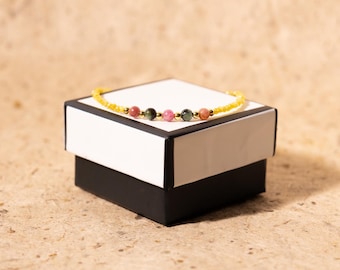May include: A delicate bracelet with a yellow beaded chain and colorful gemstone accents. The bracelet features pink, green, and orange stones, resting on a white and black box. The background is a neutral tan color.