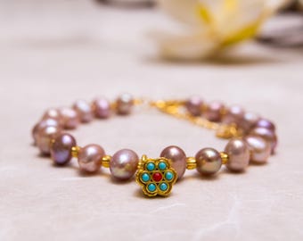 May include: A pearl bracelet featuring a gold flower charm with turquoise and red accents. The bracelet is made of light purple and gold pearls. The jewelry is displayed on a light-colored surface.