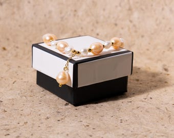May include: A pearl necklace with peach and white pearls rests on a black and white gift box. The necklace has a gold chain and a single teardrop-shaped pearl pendant. The box is on a textured, light brown surface.