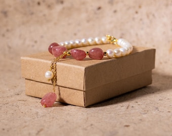May include: A beaded bracelet featuring alternating pink gemstones and white pearls, connected by gold-colored accents. A dangling chain with a pink gemstone and pearl hangs from the bracelet. The bracelet rests on a small, brown cardboard box.