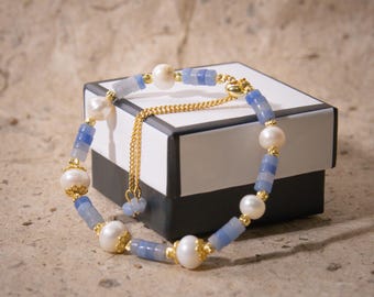 May include: A delicate bracelet featuring alternating blue and gold beads with large white pearls. The bracelet is accented with gold-colored metal and a delicate chain. The jewelry is displayed on a black and white box.