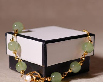 May include: A gold-toned bracelet featuring pale green, round beads. The bracelet has a delicate chain and a decorative clasp with a white flower. The bracelet is displayed on a white and black box.