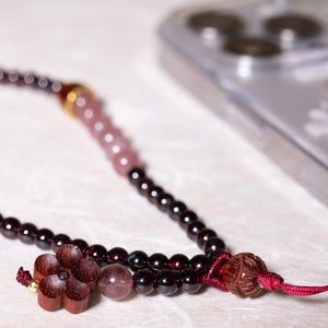 Op de afbeelding: Een donkerrode en roze kralen telefoonbedel met een houten bloemenbedel en een rood koord.