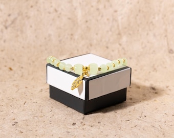 May include: A light green beaded bracelet with gold accents and a gold leaf charm rests on a black and white gift box. The bracelet features round and square beads. The background is a textured, neutral color.