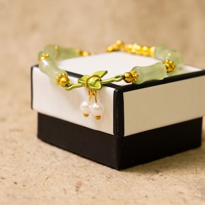 May include: A jade bracelet with gold accents rests on a white and black gift box. The bracelet features light green jade beads and gold-colored details. Two small white pearls dangle from a gold-colored clasp.