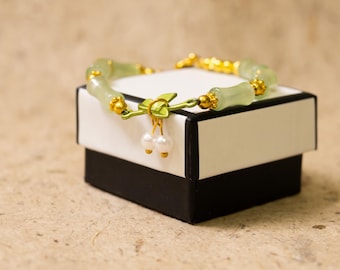 May include: A jade bracelet with gold accents rests on a white and black gift box. The bracelet features light green jade beads and gold-colored details. Two small white pearls dangle from a gold-colored clasp.