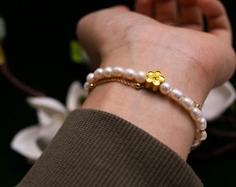 May include: A delicate pearl bracelet with a gold flower charm. The bracelet features multiple strands of white pearls and a small gold chain. The flower charm is a bright gold color. The bracelet is on a wrist.