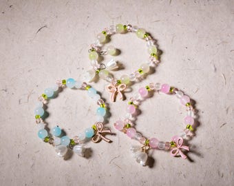 May include: Three beaded bracelets in pastel colors: light blue, pink, and yellow-green. Each bracelet features round beads, small green beads, and a charm. Charms include a pink bow and white flower-shaped pendants. The bracelets are on a textured, off-white surface.