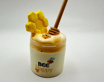 May include: A clear jar filled with honey-colored slime, topped with a yellow honeycomb and a wooden honey dipper. The jar has a white label with the text "Bee Happy" and a cartoon bee.