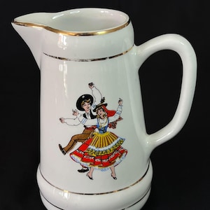 Könnte beinhalten: Ein weißer Keramikkrug mit goldfarbenem Rand und Boden. Auf der Vorderseite des Krugs befindet sich eine farbenfrohe Illustration von zwei tanzenden Figuren in traditioneller Volkstracht, ein Mann mit schwarzem Hut und eine Frau mit rotem Rock.