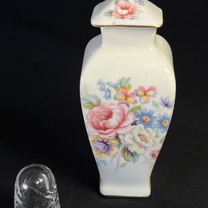 May include: A white porcelain jar with a removable lid featuring a gold-tone finial and a colourful floral pattern of pink roses, blue bellflowers, and yellow pansies. Beside the jar sits a clear, textured glass thimble with a floral motif.