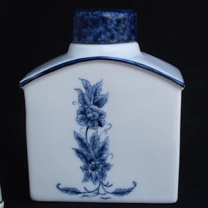 Puede incluir: Un bote de té de cerámica blanca con un patrón floral azul y una tapa azul a juego, acompañado de un pequeño dedal blanco con un delicado diseño floral rosa y verde con un borde de color dorado en la base del dedal.