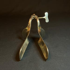Puede incluir: Un espéculo médico de metal vintage con dos hojas curvas y pulidas conectadas por un mecanismo de bisagra. Un tornillo de ajuste roscado con un mango plano en forma de T se encuentra en la parte superior para controlar la apertura de las hojas.