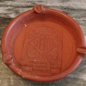 May include: A terracotta-colored ashtray with three rests. The center features a detailed embossed crest and text. A white thimble rests on the side. The ashtray is round and has a slightly raised rim.