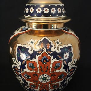 Op de afbeelding: Een keramische gemberpot met een metallic gouden afwerking, voorzien van een levendig bloemmotief in diepblauw, oranje, wit en groen. De pot heeft een bijpassend deksel met een gouden knop, versierd met een herhalend wit bloemmotief op een donkerblauwe band.