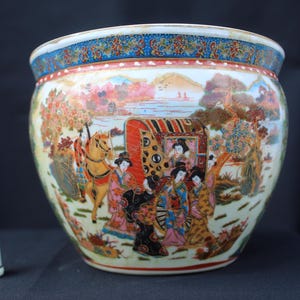 May include: A large, round ceramic bowl featuring a colorful hand-painted scene of figures, a horse, and a carriage in a landscape. The rim has a blue and gold patterned border. A small white thimble with a floral design sits to the left for scale.