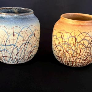 Könnte beinhalten: Zwei abgerundete Keramikvasen mit einem handgemalten Muster aus braunen Rohrkolben und blauen grasartigen Strichen. Die linke Vase hat einen blau-cremefarbenen Verlauf, die rechte einen orange-cremefarbenen. Ein kleiner Fingerhut aus klarem Glas.