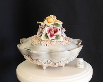 Op de afbeelding: Een decoratieve keramische serveerschaal met deksel, versierd met gebeeldhouwde rozen in roze, geel en paars. De schaal heeft een lichtblauwgrijze basis met bloemendetails en staat op drie kleine voetjes. Een bijpassende vingerhoed staat ernaast.