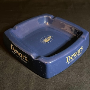 Könnte beinhalten: Ein quadratischer, dunkelblauer Aschenbecher aus Keramik mit dem goldenen Schriftzug Dewar's Fine Scotch Whisky auf zwei Seiten. In der Mitte des Aschenbechers befindet sich ein kleines, kreisförmiges goldenes Logo einer Person in traditioneller schottischer Kleidung.