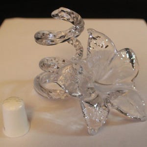 May include: A clear glass sculpture featuring a five-petaled flower attached to a spiralling, coiled stem. Beside the glass flower sits a small, solid white thimble. The glass is transparent with a smooth, reflective surface and intricate detailing.