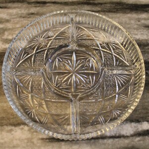 Op de afbeelding: Een heldere, ronde serveerschaal met ingewikkelde geslepen glaspatronen. De schaal is verdeeld in vier secties met een centraal sterpatroon. De rand heeft een getextureerd patroon. Perfect voor het serveren van snacks. Diameter ongeveer 25 cm.