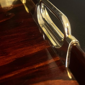 Peut inclure: Un plateau de service rectangulaire doté d'une surface en bois brun foncé poli avec une finition très brillante. Le plateau est équipé de poignées métalliques argentées présentant un design incurvé et lisse fixé sur les côtés de la base en bois.