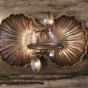Op de afbeelding: Een zilveren serveerschaal in de vorm van twee schelpen met een decoratief metalen standaard in het midden. De standaard houdt vier lepels.