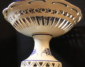 May include: A white ceramic pedestal fruit bowl with blue floral and geometric designs. The bowl has a scalloped rim with cut-out details and a pedestal base. The bowl is approximately 10 inches in diameter and 8 inches tall.