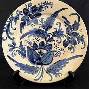 Puede incluir: Un plato redondo de cerámica con una base de color crema que presenta motivos florales de color azul oscuro pintados a mano. El diseño incluye flores estilizadas, hojas y enredaderas que se extienden por la superficie. Sobre un soporte negro.