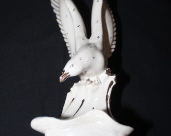 May include: A white ceramic figurine of an eagle with its wings spread wide in a soaring position. The bird features small black spots across its wings and body, with a metallic gold-toned beak and dark accents on the base it is perched upon.