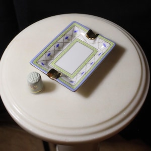 May include: A rectangular ashtray with a white centre, bordered by green and blue geometric patterns, and gold accents. A small, grey thimble with gold detailing sits beside the ashtray. The items are on a white, round surface.