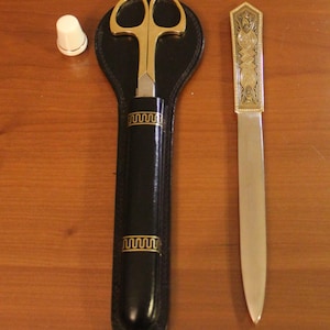 Könnte beinhalten: Ein Vintage-Schreibtischset mit goldfarbener Schere in einer schwarzen Lederscheide, einem Brieföffner mit aufwendigen Golddetails und einem weißen Fingerhut. Schere und Brieföffner haben ein klassisches Design.