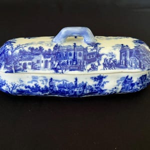 Pode incluir: Uma manteigueira de cerâmica de formato oblongo com uma tampa apresentando um padrão de transferência em azul e branco. O design retrata uma paisagem detalhada com edifícios clássicos, árvores e figuras. Uma pequena alça arqueada fica no topo.
