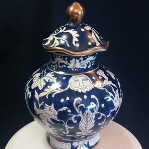 Può includere: Un vaso in ceramica blu e bianco con coperchio, con un pomello dorato. Il vaso è decorato con disegni floreali e di volti bianchi. Il bordo e la base hanno accenti dorati. Il vaso è su una superficie bianca.